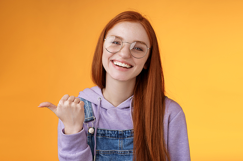 Cute helpful friendly-looking joyful european redhead woman show thumb left smiling delighted laughing pointing where find awesome store telling about interesting new promo offer, orange background.