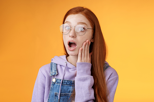 Shocked impressed speechless young redhead sensitive european girl glasses wearing hoodie dungarees drop jaw gasping astonished look left wide eyes amazed standing orange background.