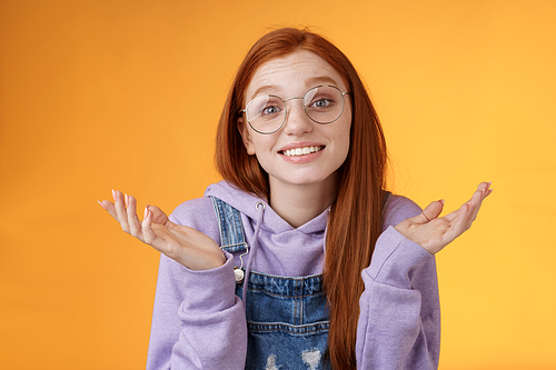 Well no big deal. Attractive silly redhead hipster girl wearing glasses smiling awkward unaware spread hands sideways shrugging confused grinning questioned uncertain what tell, orange background.