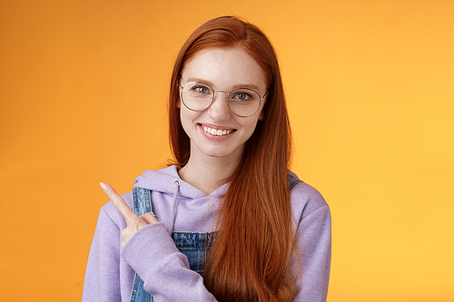 Friendly good-looking modern redhead young girl pointing left index finger showing awesome place suggesting go hang out smiling joyfully casually talking discuss new product, orange background.