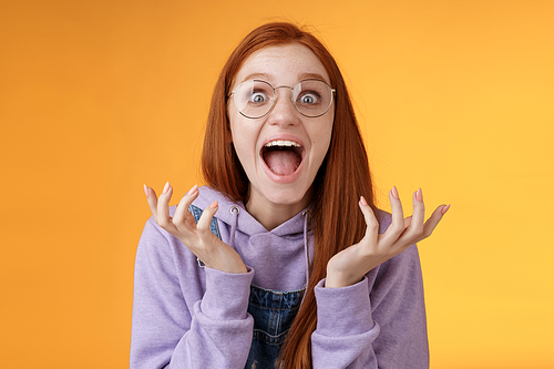 Shocked excited overwhelmed young screaming happy redhead girl wearing glasses winning hear excellent news yelling out loud rejoicing unbelievable luck raising hands spread stare surprised.