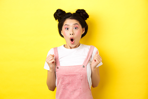 Excited and surprised asian girl clench fists, looking hopeful and amazed, say wow, standing on yellow background.