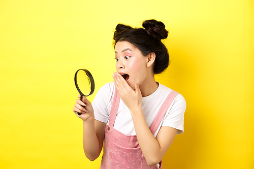 Excited silly asian girl gasping wondered, found something interesting, looking through magnifying glass, standing on yellow background.