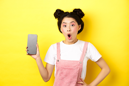 Online shopping concept. Excited stylish asian girl say wow, showing empty cell phone screen and look amazed, yellow background.