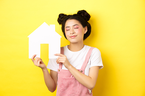 Real estate and family concept. Dreamy smiling asian woman with bright makeup, showing paper house cutout with closed eyes, daydreaming about buying property, yellow background.