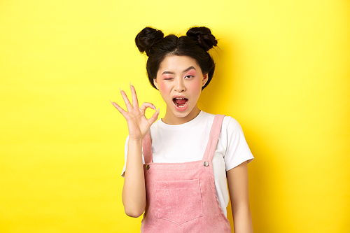 Alright. Confident asian female model with bright makeup and summer clothes, showing okay sign, winking and say yes, agree and approve, yellow background.