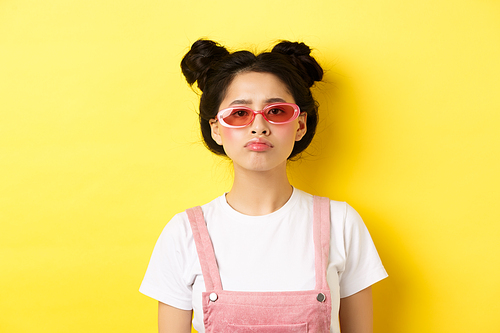 Summer and fashion concept. Bored asian teen girl in sunglasses pouting, standing moody on yellow background.