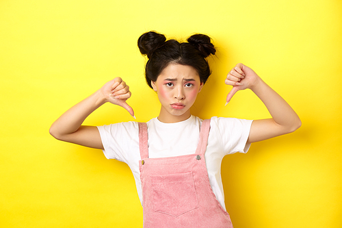 Summer lifestyle concept. Sad and gloomy asian woman with makeup, pointing fingers down and frowning upset, disappointed with unfair situation, yellow background.