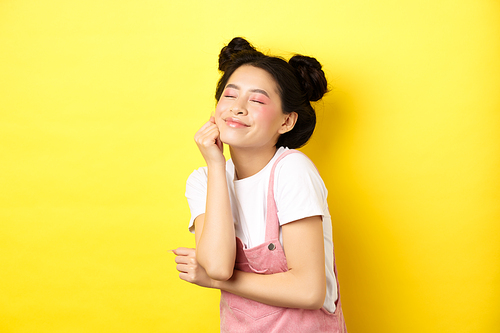 Beautiful happy woman with bright pink makeup, close eyes and touch soft skin, smiling excited, standing in summer clothes on yellow background.