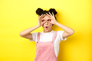 Excited asian teen girl look through hand binoculars aside, say wow and stare surprised, checking out advertisement, yellow background.
