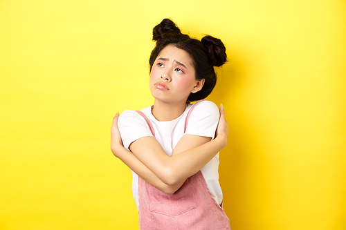 Sad lonely asian girl looking aside with upset and gloomy face, hugging herself, dreaming of love and boyfriend, yellow background.