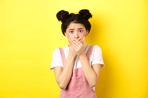 Worried asian teen girl covering mouth with hands, looking concerned and anxious, standing scared on yellow background with glam makeup and summer clothes.