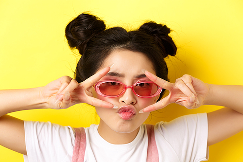Summer fashion concept. Headshot portrait of stylish girl on vacation, wearing sunglasses and showing v-signs on eyes, pucker lips cool, standing on yellow background.