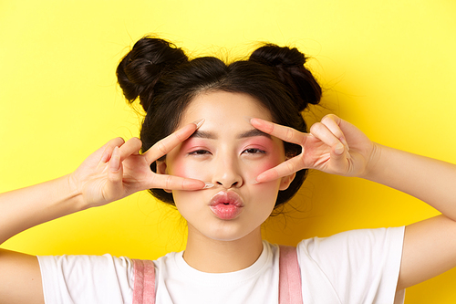 Carefree asian girl showing v-signs and kissing lips, standing on yellow background.