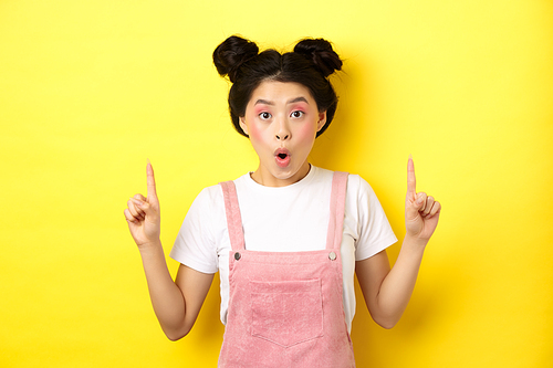 Excited asian female model with glamour makeup, pointing fingers up and say wow amazed, checking out promo deal, standing on yellow background.