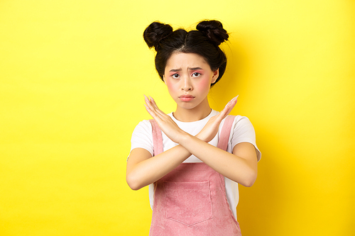 Sad girl begging to stop, frowning uspet and showing cross sign, say no, standing gloomy on yellow background.