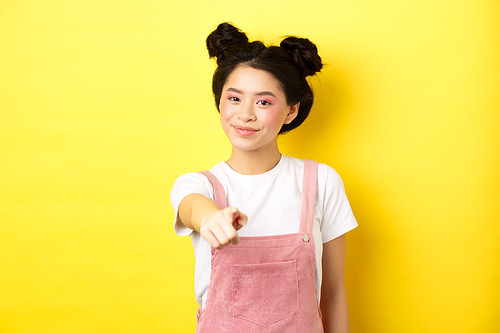 Stylish japanese girl with beauty makeup pointing at camera, smiling at you, beckon and invite people, standing on yellow background.