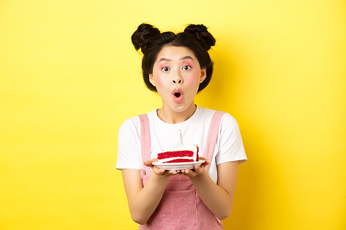 Holidays and celebration. Excited asian birthday girl blowing candle on party cake, making wish on b-day, standing against yellow background.