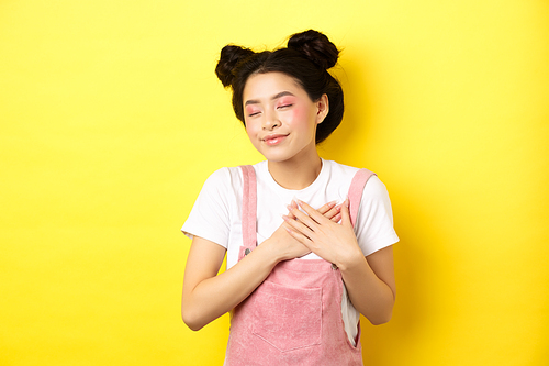 Romantic asian girl with makeup, wearing summer clothes, holding hands on heart and smiling tenderly, remember tender memory, standing on yellow background.