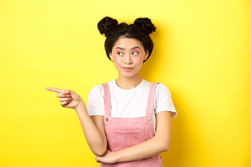 Cute beauty girl with makeup and summer clothes, pointing and looking left at promo banner, standing curious on yellow background.