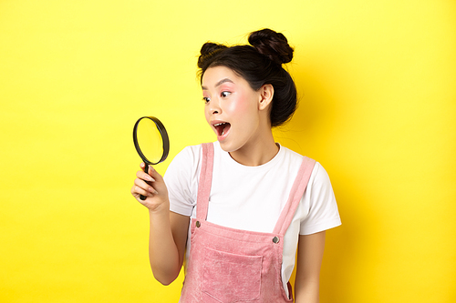 Excited asian girl found interesting thing, looking through magnifying glass amazed, standing on yellow summer background.
