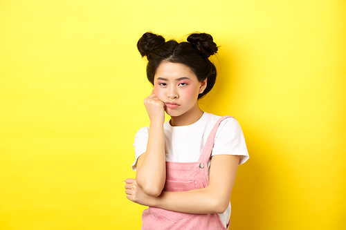 Bored asian teen girl with stylish makeup and summer clothes, looking reluctant and indifferent, standing on yellow background.