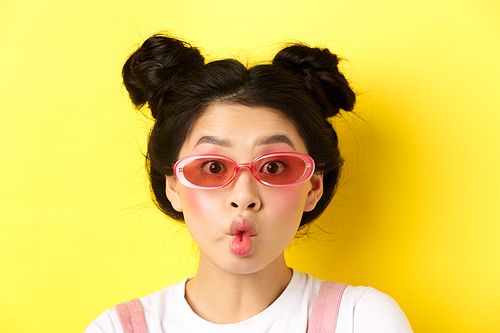 Summer fashion concept. Silly glamour girl in sunglasses, pucker lips like fish and looking funny at camera, standing against yellow background.