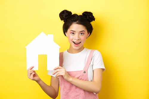 Real estate and family concept. Cute asian woman with bright makeup and stylish hairbuns, showing paper house cutout, smiling determined, yellow background.