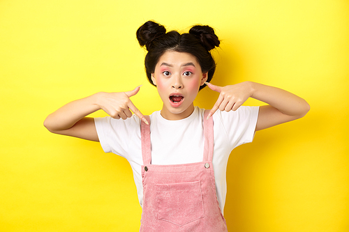 Excited teenage asian girl with beauty makeup, pointing fingers down and open mouth fascinated, standing happy on yellow background.