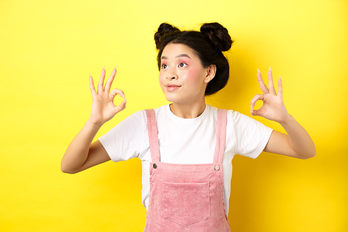 Romantic and beautiful asian girl looking at logo left, showing okay signs in approval, standing on yellow background.