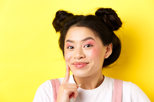 Beauty. Stylish asian girl with bright glamour makeup, touching soft and shiny facial skin, smiling happy at camera, standing on yellow background.