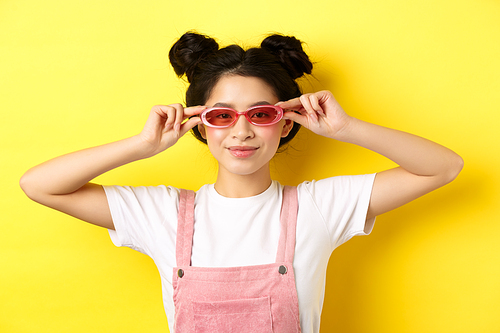 Beautiful asian girl put on stylish sunglasses and smiling, wearing trendy summer outfit, standing on yellow background.