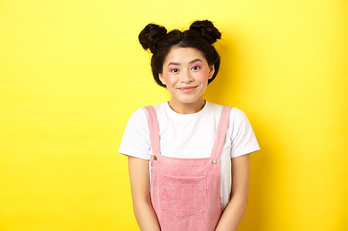 Cute asian woman with makeup and summer clothes, smiling silly and happy at camera, yellow background.
