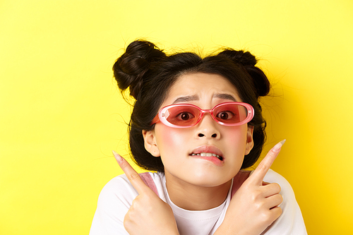 Summer fashion concept. Headshot of worried indecisive asian girl, pointing fingers sideways and showing two ways, cant choose, wearing sunglasses.