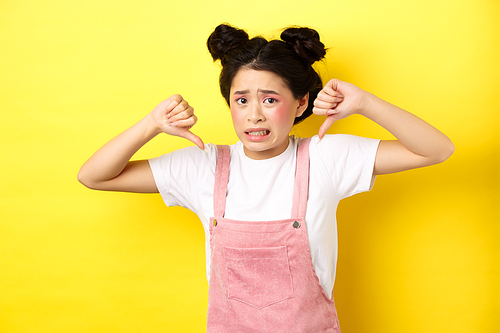 Disgusted asian girl showing thumbs down and cringe from bad product, standing in summer clothes against pink background.
