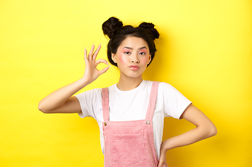 Sassy teenage asian girl with bright pink makeup, showing okay sign and nod in approval, approve product with serious face, not bad gesture, standing against yellow background.