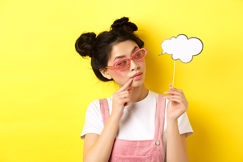 Summer and fashion concept. Stylish glamour asian girl in sunglasses, holding comment cloud party mask and looking aside, yellow background.