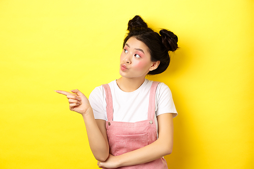 Pensive asian woman with beauty makeup, pointing and looking left with curious face, interested in advertisement, standing on yellow background.
