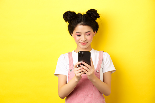 Stylish asian girl with glamour makeup using mobile phone, smiling at screen, standing on yellow background.