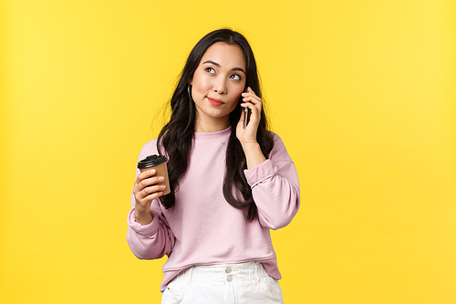 People emotions, lifestyle leisure and beauty concept. Cute stylish asian woman looking up thoughtful as talking on mobile phone and drinking coffee from takeaway cup, yellow background.