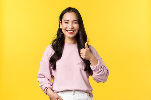 People emotions, lifestyle and fashion concept. Cheerful friendly-looking asian girl with happy smile, recommend beauty product, showing thumb-up, like and agree to something, yellow background.