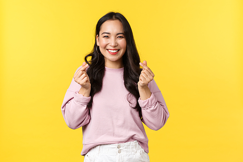 People emotions, lifestyle and fashion concept. Kawaii asian girl with beautiful smile showing korean hearts gesture sending positive and joyful vibes, standing yellow advertisement background.