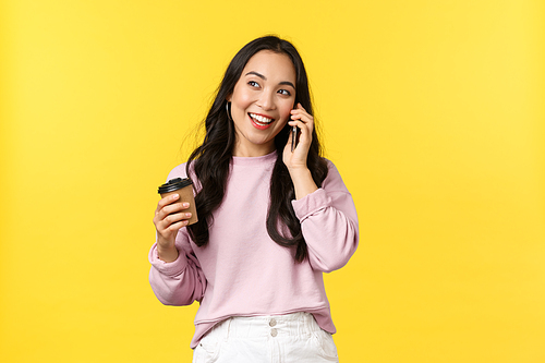 People emotions, lifestyle leisure and beauty concept. Modern cute asian girl talking on phone with friend and smiling, drinking coffee casually, hold takeaway cup, standing yellow background.