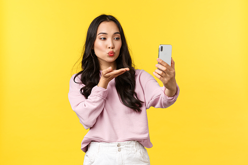 People emotions, lifestyle leisure and beauty concept. Stylish and flirty asian girl taking photo for social media, make selfie, sending air kiss at smartphone camera, stand yellow background.