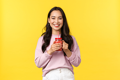 People emotions, lifestyle leisure and beauty concept. Cheerful smiling asian girl enjoying fresh smoothie, drinking juice and looking camera enthusiastic, standing yellow background.