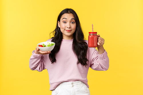 People emotions, healthy lifestyle and food concept. Silly and cute asian beautiful girl showing smoothie and vegan salad, smiling as trying stay on diet, standing yellow background.