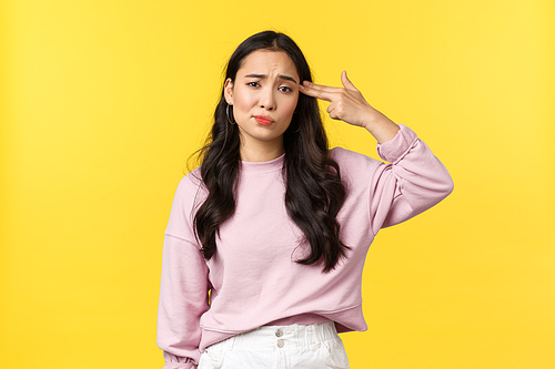 People emotions, lifestyle and fashion concept. Bothered and annoyed asian girl making fake gun over temple as if shooting herself from irritation or distress, standing gloomy yellow background.