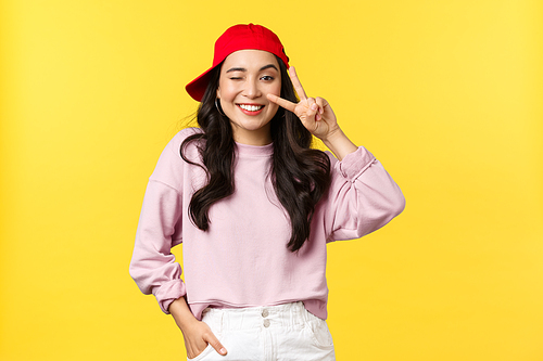 People emotions, lifestyle leisure and beauty concept. Cheerful teenage asian girl in hip hop red cap, wink and smiling broadly, showing peace sign, standing yellow background.