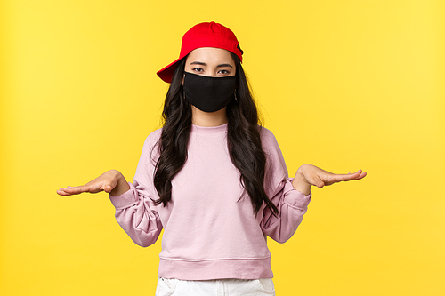 Covid-19, social-distancing lifestyle, prevent virus spread concept. Unbothered young asian girl in face mask and red cap staying low, showing something little, yellow background.