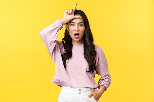 Lifestyle, emotions and advertisement concept. Arrogant and sassy confident young asian woman mocking person for losing, showing loser sign on forehead, stand yellow background.